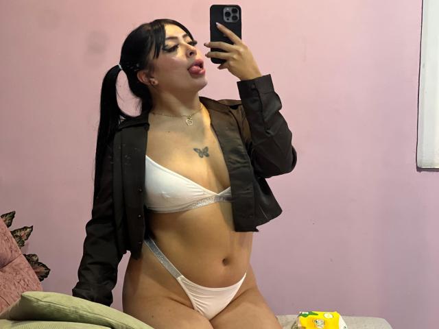 KateWelsin with pigtails, tongue out, wearing a black jacket, white bikini, showing her tits and pussy, taking a selfie with a black phone, pink wall background.