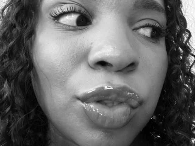 Het gezicht van LisaMendez op een zwart-wit foto, waarbij ze naar de zijkant kijkt met glanzende lippen, krullend haar en licht gefronste wenkbrauwen. Haar glanzende, nat uitziende lippen zijn opvallend..