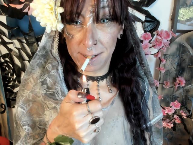 Grethel, una donna con capelli rosso scuro, un anello al naso e un piercing al setto, che fuma una sigaretta, indossa un velo di pizzo, una collana nera e un copricapo floreale, tenendo un accendino nella mano destra..