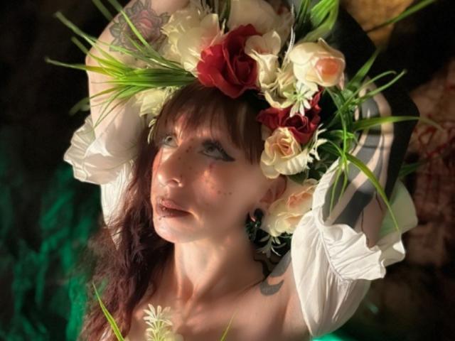 GrethelMulher with a floral headpiece, white puffed sleeves, and green leaves, has pale skin, brown hair, and tattoos, looking up with makeup and a rose on her chest.