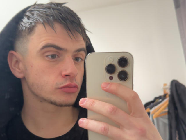 LorenzoZo, com cabelo preto e molhado, pele clara e uma barba discreta, lábios curvados para baixo, segurando um iPhone dourado, tirando uma selfie com a mão direita..