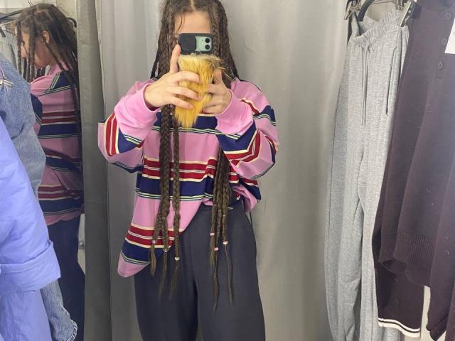 HotCaramelsDuo with long brown dreadlocks, pink and blue striped shirt, black pants, holding phone, mirror reflection, grey curtain, and hanging clothes, showing yellow hair in phone's front.