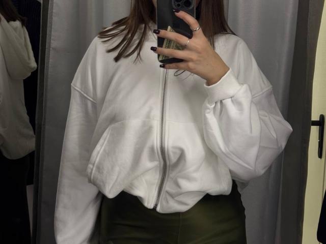 HotCaramelsDuo wearing a white zip-up hoodie, green pants, black nails, holding a phone in front of their face, reflection in mirror, visible ring on left hand.