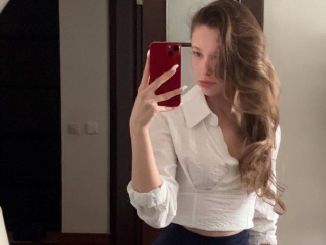 Eira Kilian, met lang, golvend bruin haar, een wit overhemd en donkerblauwe broek, houdt een rode telefoon vast en maakt een spiegelzelfportret, waarbij haar borsten en billen zichtbaar zijn..