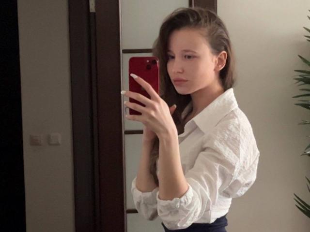 Eira Kilian mit langen, braunen Haaren, einem weißen Hemd und einer schwarzen Hose, die ein rotes Telefon hält, macht ein Selfie im Spiegel und zeigt dabei ihre schlanken Finger und ihr dezentes Lächeln..
