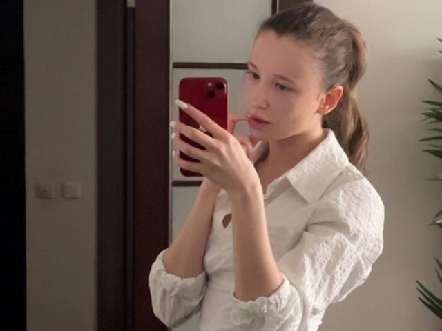 Eira Kilian met een paardenstaart, een wit overhemd met knopen, die een rode telefoon vasthoudt, een spiegel-selfie maakt, er licht geïrriteerd uitziet, en haar kleine borsten en slanke billen laat zien..