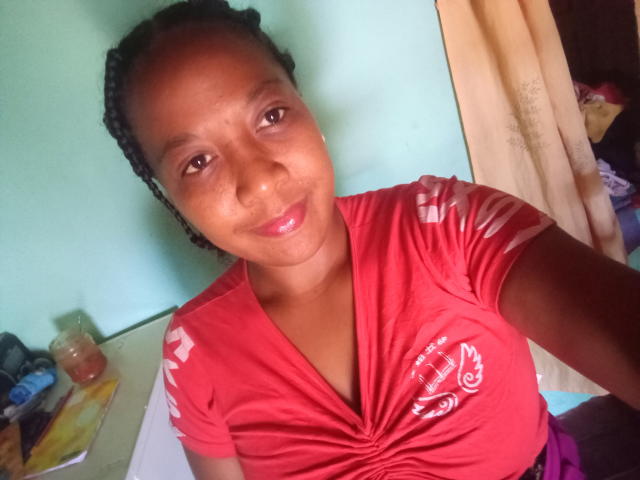 MileyHot69, avec une peau brune, un petit nez et des tresses bouclées, portant une chemise rouge avec des motifs blancs, souriant à la caméra, montrant ses seins, dans une pièce très éclairée..