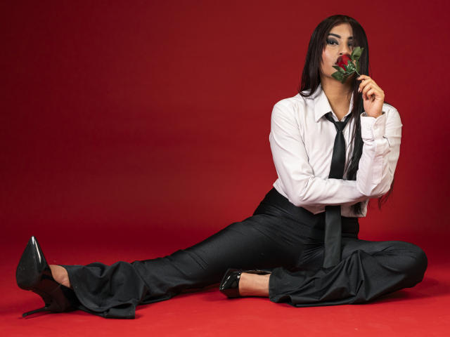 EmmaaRodriguez with long black hair, white shirt, black tie, and pants, sitting on red floor, holding rose to her mouth, wearing black high heels, showing her ass.