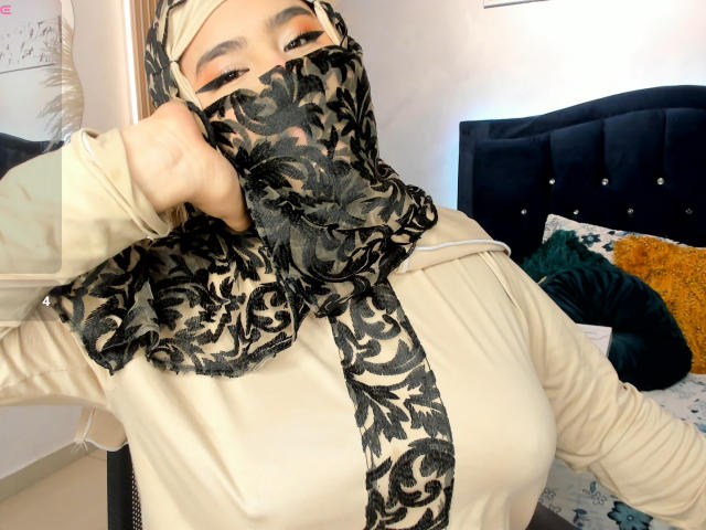 DaliilaAli with black floral mask, beige headscarf, and beige long-sleeve shirt, adjusting mask, revealing orange eyeshadow, large tits, and white wall in background.