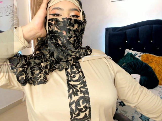 DaliilaAli with black floral mask, beige headscarf, and cream shirt, showing off her tits and holding her headscarf up. Bedroom with dark blue headboard and yellow pillow.