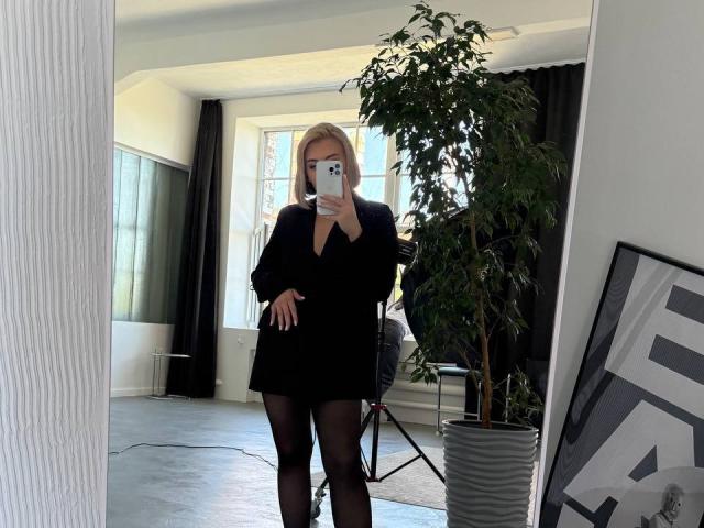 SassyJam, con un corte de pelo rubio, un vestido negro y medias negras, se está tomando un selfie frente al espejo, mostrando su lindo trasero, de pie frente a una planta, usando un teléfono azul..