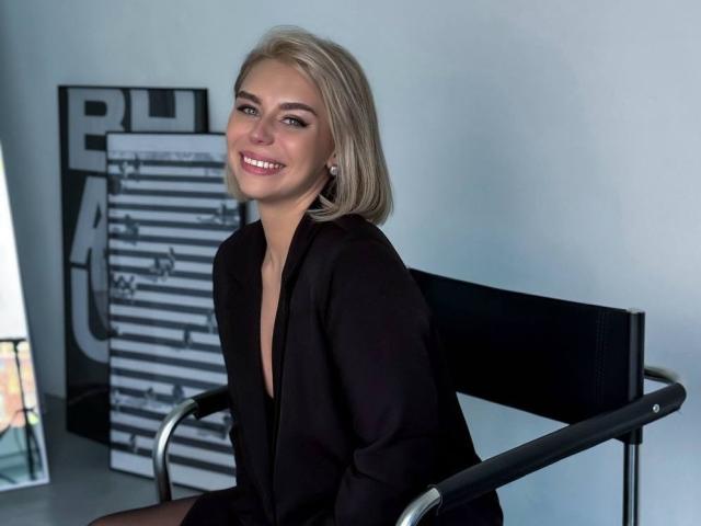 SassyJam is a blonde woman with a shoulder-length bob, smiling at the camera. She wears a black blazer over a deep V-neck. She's seated on a black metal chair with a backrest. Her skin is fair, and she has light makeup. The background has a vertical black and white sign with text and abstract shapes. The image is sharp with even lighting. SassyJam's expression is friendly and confident.