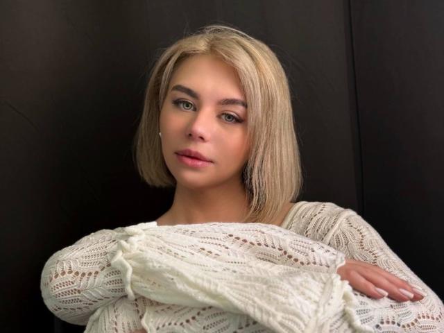 SassyJam with blonde bob, green eyes, and white knit sweater, arms crossed, looking at camera, slight smirk, soft lighting, dark background.