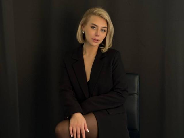 SassyJam, rubia, vestida con un traje negro, sentada con las piernas cruzadas, con las manos sobre la rodilla, mirando ligeramente hacia un lado, con una sonrisa sutil y segura, en un entorno oscuro y minimalista..