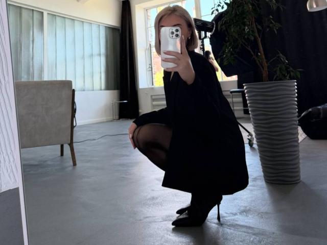 SassyJam squats in black dress, sheer stockings, and heels, holding phone to mirror, showing her ass, with blonde hair, in a room with a potted plant.
