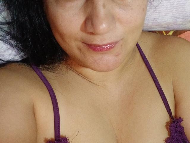 KarinaCecillia's face with shiny pink lips, dark hair, and slight smile, wearing a purple lace bra, showing her bare chest and small tits. Her skin is light brown.