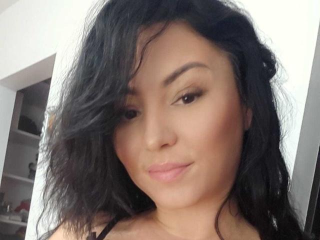 AmelyBrown with wavy black hair, brown eyes, and a subtle smile, showing her cute face and glossy pink lips. Her ass is not visible, just her upper body.