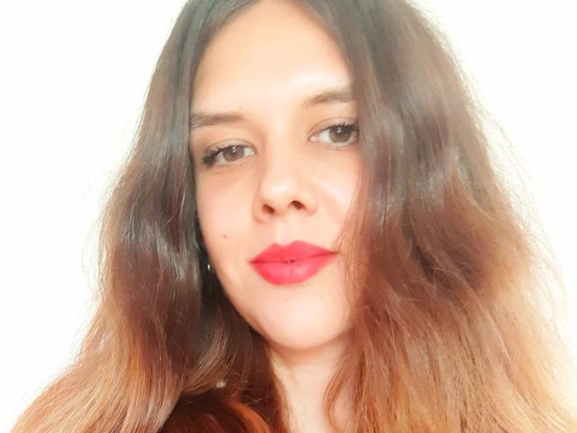 SuperLongLegs69 has light brown wavy hair, red lipstick, and a small mole on her left cheek. She's looking slightly to the side with a subtle, confident smile.