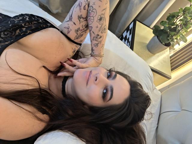 ChristineBray with large tits, lying on a bed, wearing a black lace top, choker, and tattooed arm, looking at the camera with a slight smile.