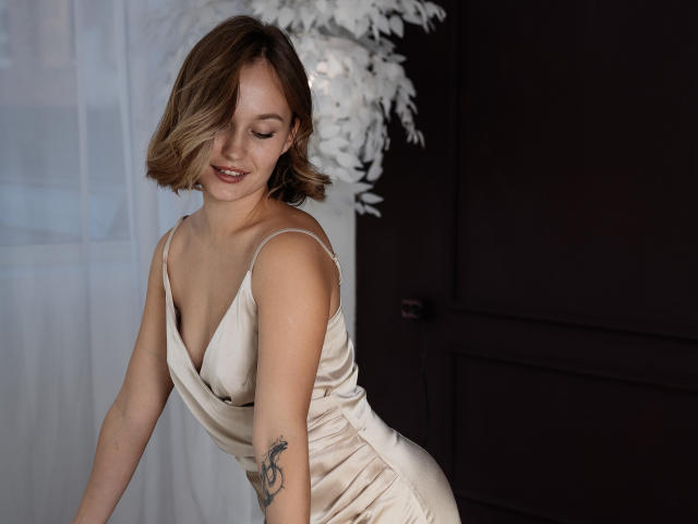 BrittanySweet in a silky cream dress, showing her small tits and tattoo on her right arm, with shoulder-length blonde hair, smiling softly, leaning forward.