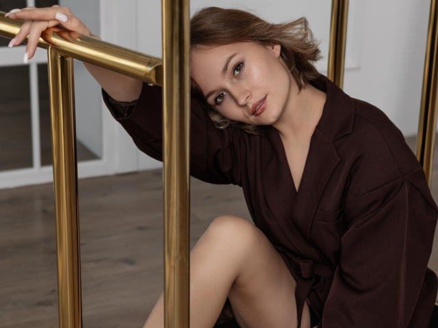 BrittanySweet with light brown hair, wearing a brown dress, leaning on gold railings, looking at camera with a soft, sultry expression, showing a bit of her bare thigh.