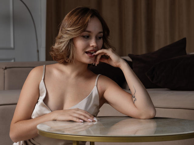 BrittanySweet with blonde hair, wearing a silky white top, sitting at a table, her hand near her mouth, showing a tattoo on her right arm, and a cute smile.
