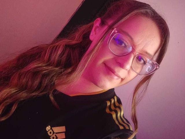 CleareRems with wavy brown hair, clear glasses, and a black Adidas shirt with yellow stripes, smiling slightly. Pink and purple lighting. Boobs not visible.