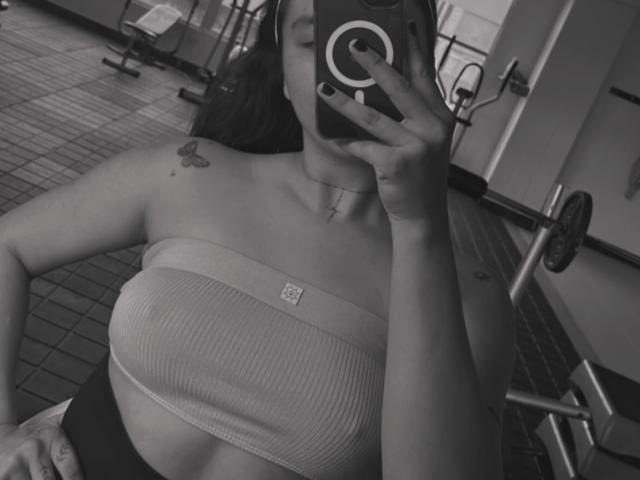 Elizabeth Macy prenant un selfie dans une salle de sport, portant un débardeur côtelé qui expose ses seins, avec un petit tatouage de papillon sur son épaule droite, tenant un téléphone avec un anneau..