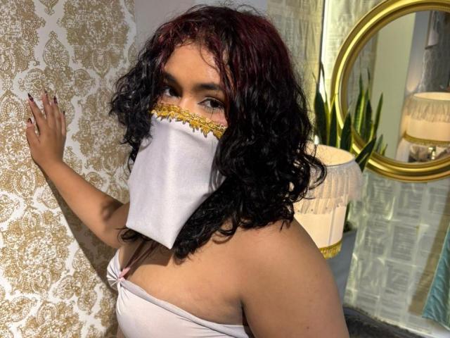 Angelitha Parker, com cabelos pretos e ruivos encaracolados, usando uma máscara branca com detalhes dourados e um top rosa sem alças, está em pé em frente a uma parede estampada, mostrando seus seios..