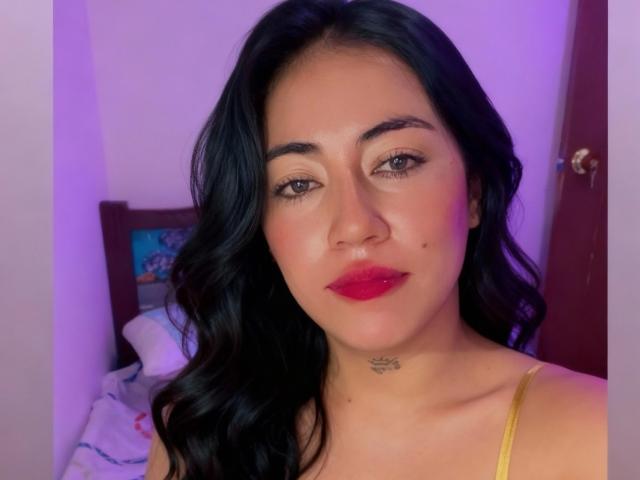 ZoeMonroeI a de longs cheveux noirs, du rouge à lèvres et un petit tatouage sur le cou. Elle porte un haut jaune, et il y a une douce lumière violette en arrière-plan..
