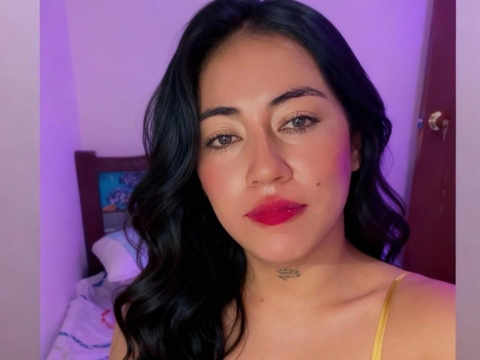 ZoeMonroeI a de longs cheveux noirs, du rouge à lèvres et un petit tatouage sur le cou. Elle porte un haut jaune, et il y a une douce lumière violette en arrière-plan..