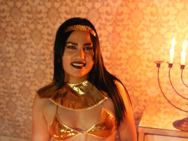 TifaniGreys with gold sequined bra, shiny gold collar, and headpiece, smiling, black hair, gold eyeliner, orange light, candelabra on right, patterned wallpaper in background.