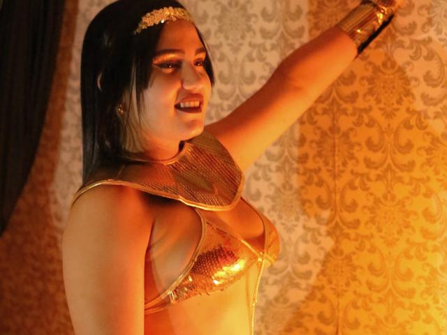 TifaniGreys has a dark, straight-haired, medium-build physique, wearing a gold, shimmering, chain-like top that reveals her medium-sized breasts. She's smiling with her right arm raised. Her face has bold makeup with dark lipstick. She wears a gold headpiece and a gold arm bracelet. The background has an ornate, patterned, golden wallpaper. The lighting is warm, highlighting her skin and the metallic texture of her top. The image has a theatrical, almost fantasy-style vibe.