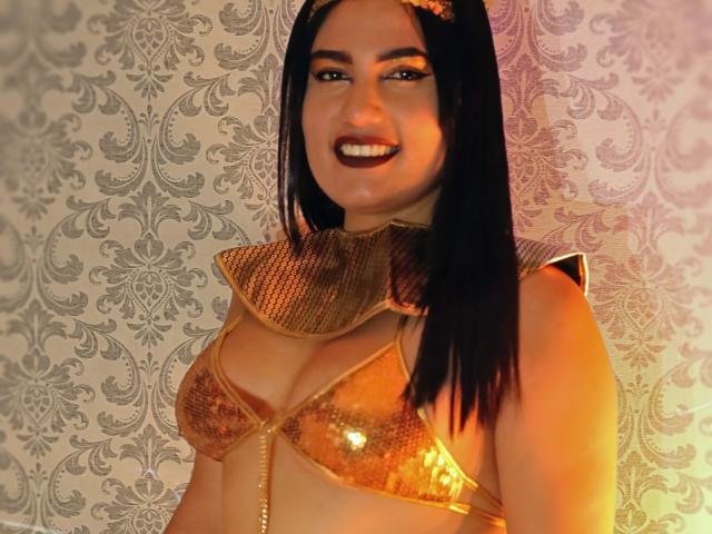 TifaniGreys stands against a patterned wallpaper. She has long black hair, dark makeup including bold red lipstick, and is smiling. She wears a shiny gold bra with sequined fabric and a metallic gold collar with chain detailing. The light has an orange-yellow hue, casting warm tones on her skin. Her bra highlights her medium-sized breasts. The background is textured with intricate patterns. The image is a photograph.