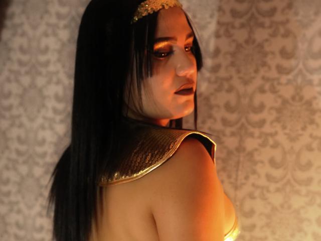 TifaniGreys has long black hair, looking over her shoulder with a serious expression. She wears a gold headpiece and a metallic shoulder piece. Her skin is warm-toned. The light casts an orange glow on her right shoulder. The background is a patterned wallpaper. Her makeup includes dark eyeliner and lipstick. The image has a slightly blurred effect, emphasizing the warm light and her expression. The metallic shoulder piece has a textured, shiny surface.
