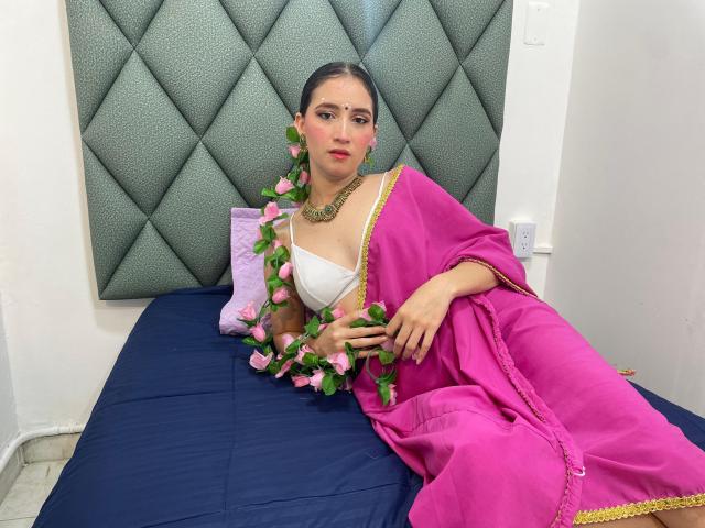 JalimaMona with dark hair, pink sari, white bra, pink flower necklace, green earrings, and pink flowers, lying on a blue bed, looking at the camera, sexy.