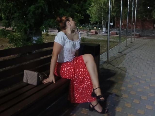 Sierens, portant une jupe à pois rouges, un haut blanc et des sandales noires, assise sur un banc en bois sombre, dos à la caméra, la tête inclinée vers le haut, la main droite sur un sac marron, dans un parc la nuit..