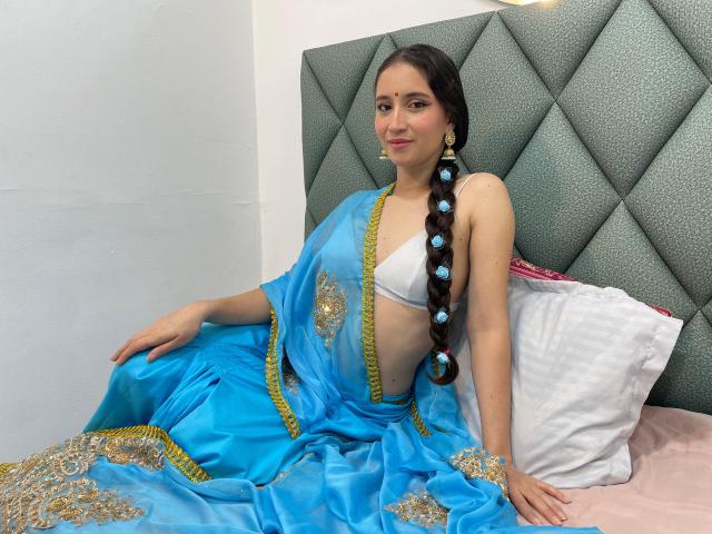 JalimaMona with long black braid, blue sari with gold embroidery, white bra, sitting on bed, grey diamond-patterned headboard, smiling, earrings, white pillow, pink bedsheet, cute.