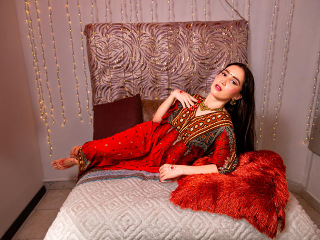 JalimaMona with red, patterned boho dress, lying on bed, legs up, arms crossed, foot adorned with henna, red lips, brown hair, jewelry, red furry blanket, white quilted bed.