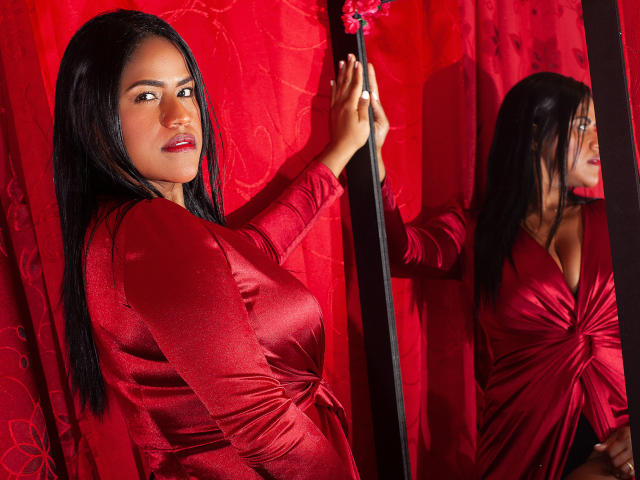 IvannaDuran with long black hair, red dress, and cleavage, stands against a red wall, touching a mirror, reflecting her sexy ass and red lips. She's looking at the camera.