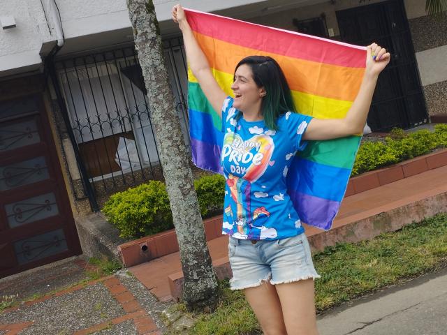 LunaVenus with green-tipped black hair, blue Pride shirt, and denim shorts, holding a rainbow flag, smiling, standing by a tree, wearing a "Pride Pro" t-shirt.