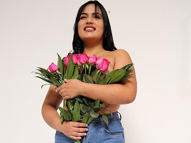 TifaniGreys com cabelo preto, sorrindo, segurando rosas cor-de-rosa, sem blusa, vestindo jeans azuis, mãos cobrindo os seios, folhas verdes, fundo branco, pele clara, fofa, expressão feliz..