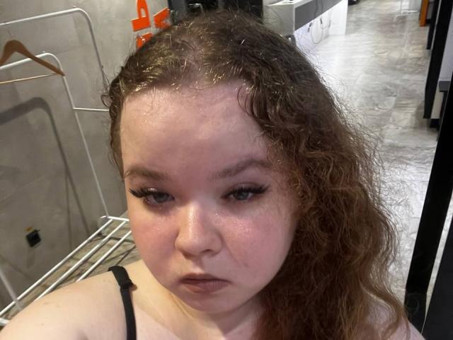 MimiEmily with curly brown hair, shiny forehead, black eyeliner, and a serious expression, wearing a black spaghetti strap top, in a room with metal and wooden hangers.