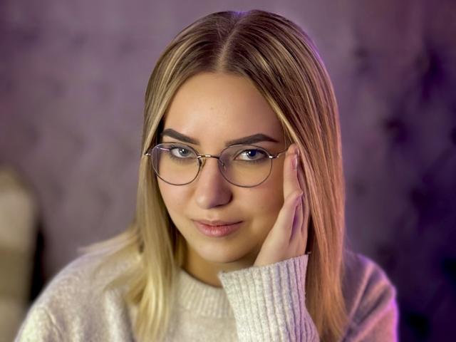 CarolFlexy with blonde hair, wearing glasses, and a white sweater, touching her face with a slight smile, blue eyes, and fair skin. She has small tits and a cute face.