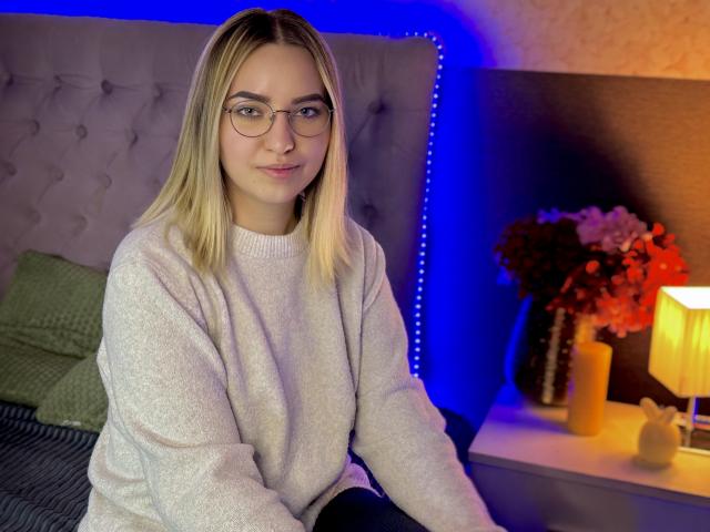 CarolFlexy with blonde hair, glasses, and a cute smile, wearing a white sweater, sitting on a bed with a gray headboard, and a yellow lamp on the right.