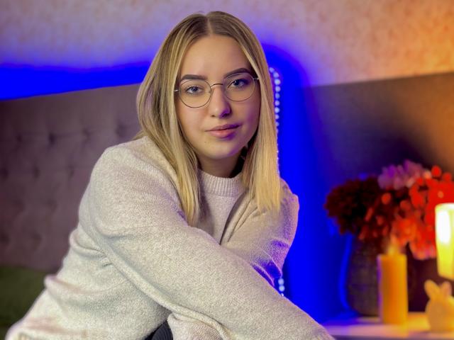CarolFlexy with blonde hair, glasses, and a grey sweater, sitting on a bed, blue light in background, looking at camera with a slight smile. Her tits are small.