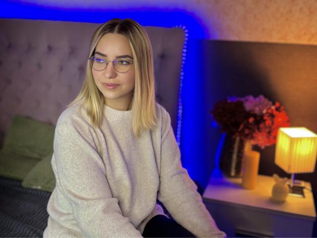 CarolFlexy with blonde hair, glasses, and a pale pink sweater, sitting on a bed, blue light in background, smiling, looking at camera, hands on black pants.