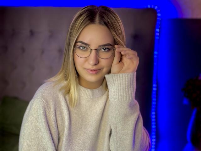 CarolFlexy with blonde hair, glasses, and a cute smile, wearing a white sweater, touching her glasses with a hand, sitting in a blue-lit room.