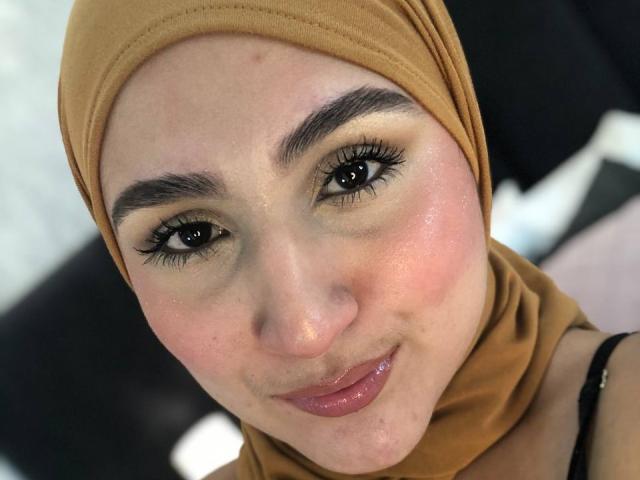 HiilaryDolce with brown headscarf, glossy pink lips, dark eyebrows, and long eyelashes, smiling softly, showing slight blush on her fair skin, and subtle freckles on her nose.
