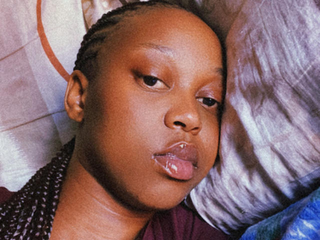HotLushiana with dark brown skin, braided hair, and her tongue slightly out, looking off to the side. Her glossy lips are slightly parted. She's lying on a pillow.